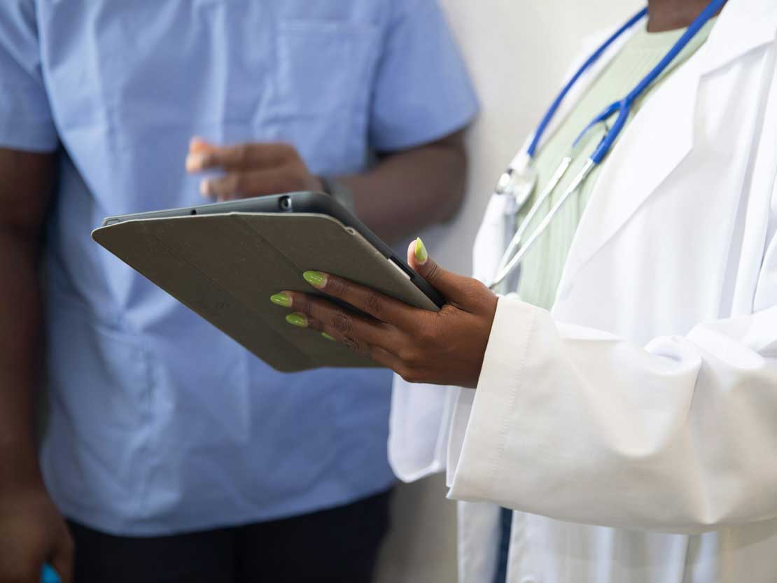 A person in a white coat and stethoscope shows their clipboard to another person wearing scrubs.