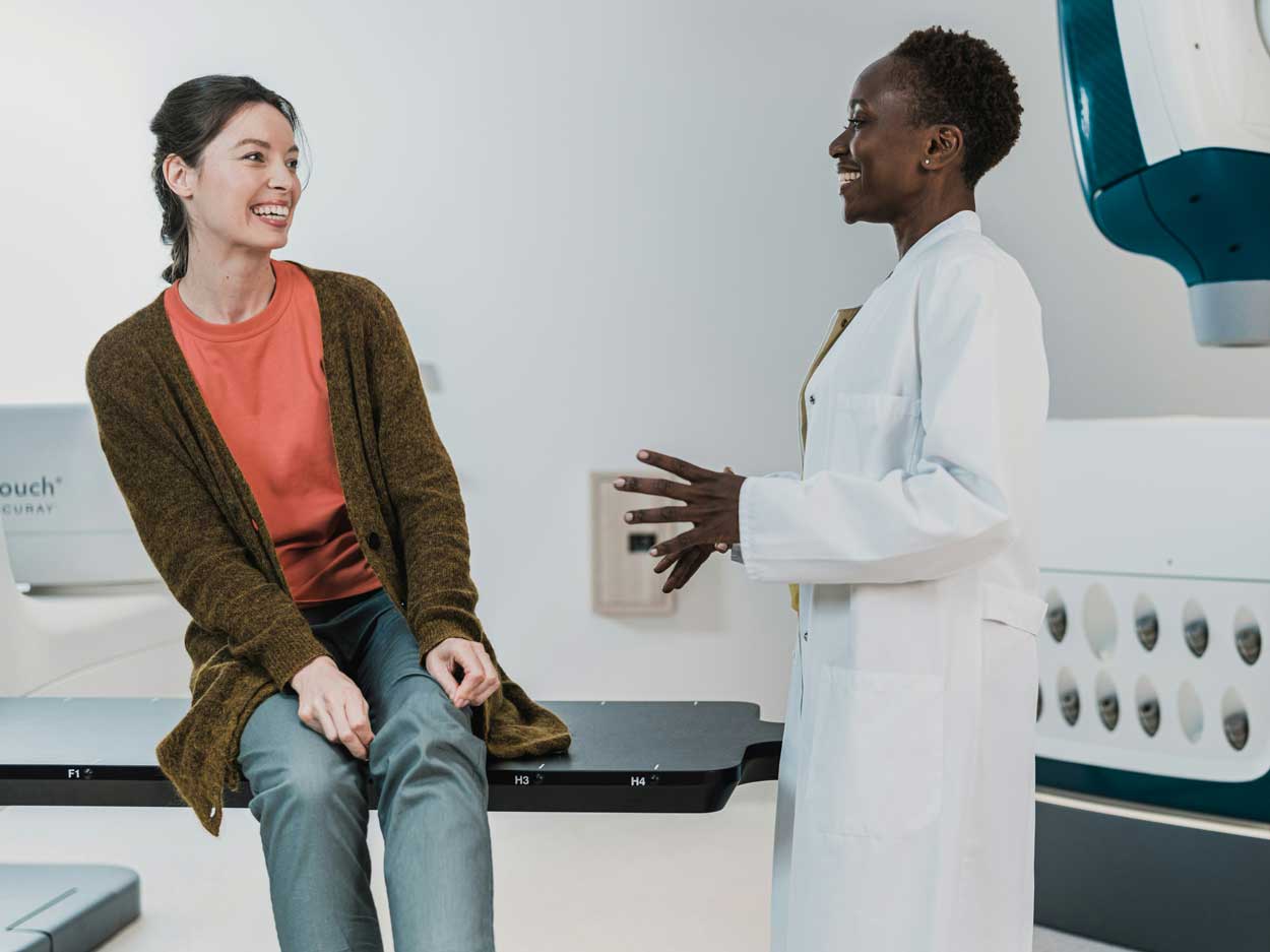 A woman in a white coat speaks with a female patient. They are both smiling.