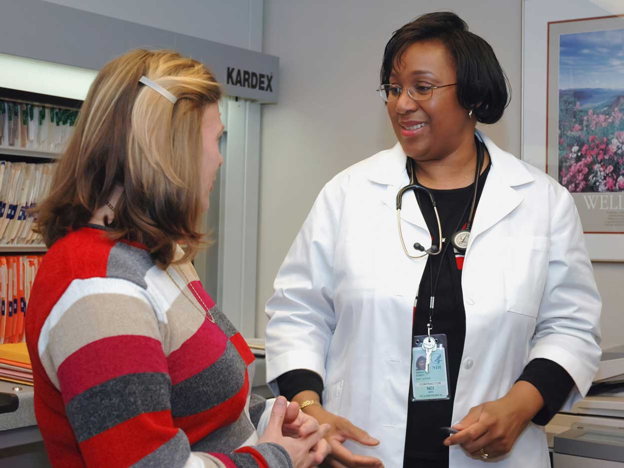A woman in a white coat and stethoscope smiles as she talks with a patient in an exam room.