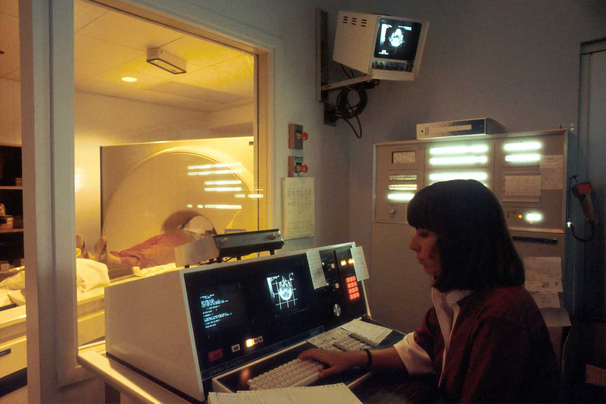 A woman watches the screen while a patient is scanned in a machine.