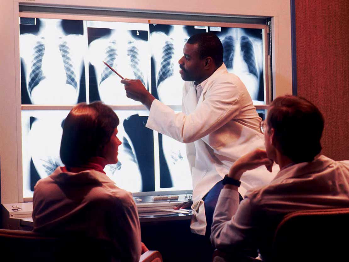 A man in a white coat discusses xrays with two other people, also in white coats.