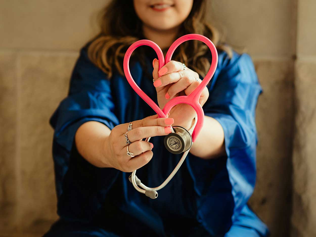 A woman in dark blue scrubs holds out a pink stethoscope in the shape of a heart.